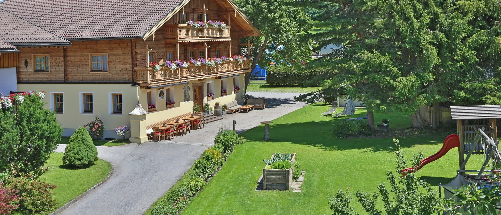 Das Bauernhaus des Kasparbauer mit Blumenbalkonen, Wiese und Bäumen vor bewaldeten Bergen